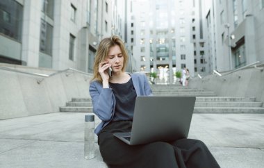 Akıllı telefondan konuşan kafası karışmış bir iş kadını ofisin yanındaki merdivenlerde laptopuna bakıyor. Kafası karışmış ofis çalışanı, iş merkezinin merdivenlerinde oturmuş bilgisayarındaki sorunlarla uğraşıyor..