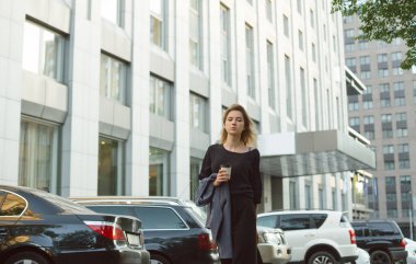 Modern şehrin caddesinde gezen, şık, sıradan giysiler içinde genç bir kızın portresi. Şehir merkezinde yürüyen genç bir kadın, ofisin ve otoparkın yakınındaki kameraya bakıyor..