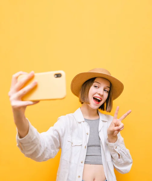 黄色の顔をした可愛い幸せな女の子の肖像画は スマートフォンで自撮りし 上に帽子とシャツを着て 平和のジェスチャーを親指を表示します 陽気な女がわがままを言う — ストック写真