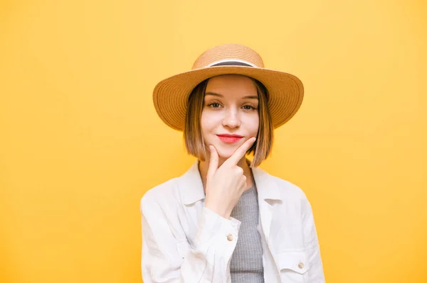 Closeup portrait of pretty lady in hat and summer clothes looking into camera with smile on face and thoughtful face. Pensive lady looking at camera on yellow background.