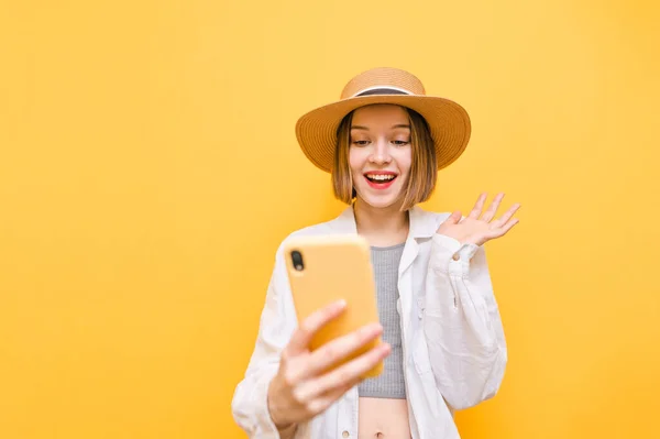 Happy lady with smartphone in hand isolated on yellow background, looking into smartphone screen with happy face and smiling. Joyful hiker girl in hat uses smartphone isolated on yellow background.