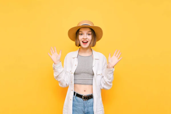 Portrait of joyful lady in light summer clothes on yellow background, looks into camera with happy face and arms raised up with joy. Shocked girl with happy face isolated on yellow, wearing sun hat.