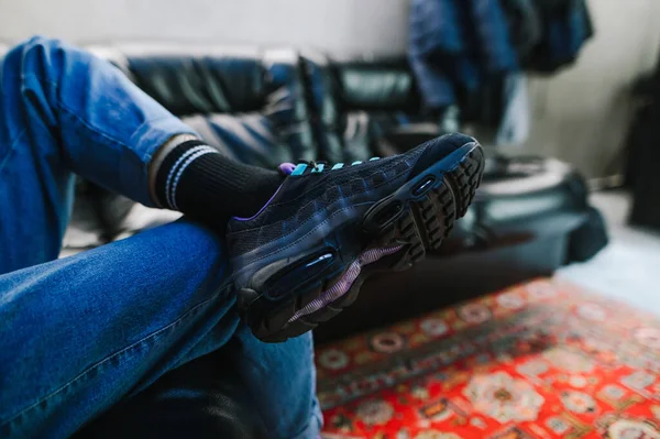 流行のスニーカー クローズアップ写真を室内の革のソファに座っている男の足は 足に彼の足を投げた ジーンズと流行のスニーカーの男は 靴に焦点を当てます — ストック写真
