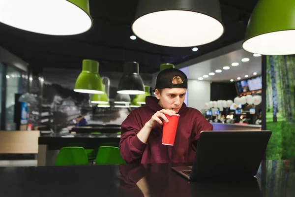 一个穿着休闲装的专注的年轻自由职业者的画像 使用笔记本电脑 在快餐店里唱一支凉爽的红可乐 聚精会神地凝视着屏幕 自由职业概念 — 图库照片