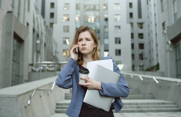 Puzzled businesswoman in front of office building having phone conversation holding laptop and documents.Female office worker with puzzled expression dealing with deadlines holding papers and computer