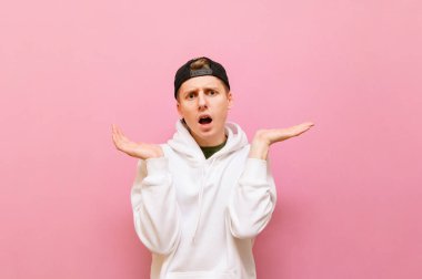 Kafası karışık genç bir adamın portresi pembe bir arka planda duruyor, kollarını yana açıyor ve şaşırmış bir yüzle kameraya bakıyor. Pembe duvarda kafası karışmış bir genç.