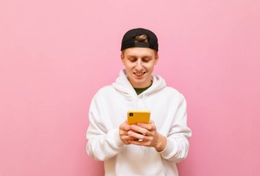 Beyaz kapüşonlu ve şapkalı neşeli genç adam portresi elinde akıllı telefonla pembe arka planda duruyor ve internet kullanıyor. Günlük giysiler içindeki genç adam akıllı telefon kullanıyor, izole..