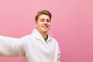 Belom giysili gülümseyen sarışın adam pembe arka planda selfie çekiyor, mutlu yüzlü kameraya bakıyor. Mutlu genç adam kamera önünde fotoğraf çekiyor uzanmış elleriyle, pembe üzerinde izole edilmiş..