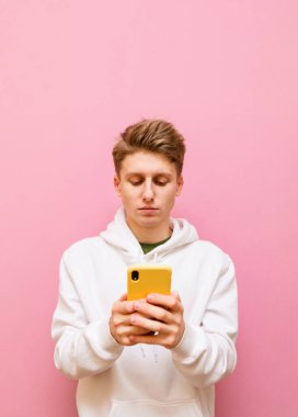Beyaz kapüşonlu, konsantre olmuş genç bir adamın portresi pembe arka planda elinde akıllı bir telefonla dikiliyor ve dikkatlice ekrana bakıyor. Öğrenci akıllı telefondan internet kullanıyor, izole edilmiş..