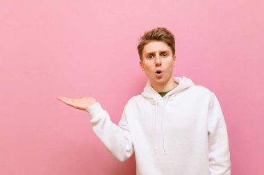 Beyaz kapüşonlu adam pembe arka planda duruyor, elinde fotokopi alanı tutuyor ve kameraya bakıyor. Şok olmuş genç adam elini bir kenara kaldırdı ve uzaklara baktı..