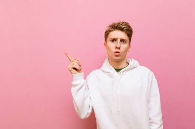 Pembe arka planda izole edilmiş beyaz giysili, şaşırmış genç bir adamın portresi, şok olmuş yüzüyle kameraya bakıyor ve boş alanı işaret ediyor. Boşluğu kopyala