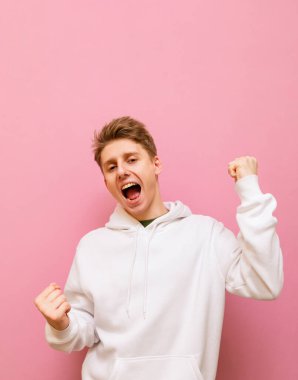Hafif günlük giysiler içinde neşeli bir adamın portresi pembe arka planda zafere seviniyor, kameraya bakıyor ve elleri havada gülümsüyor. Dikey fotoğraf. Boşluğu kopyala