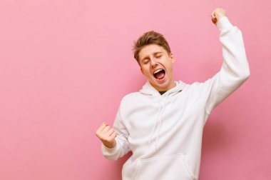 Beyaz kapüşonlu neşeli genç adam pembe arka planda mutlulukla bağırıyor. Yakışıklı, duygusal olarak mutlu, izole. Öğrenci sınavı geçti. Boşluğu kopyala