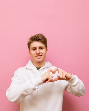 Pembe arka planda izole edilmiş beyaz kapüşonlu, gülümseyen genç bir adam, yüzünde gülümsemeyle kameraya bakıyor ve kalp hareketi sergiliyor. Adam pembe arka planda aşkının jestini gösteriyor..