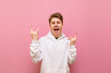 Beyaz kapüşonlu oyuncu genç adam pembe arka planda çiğnenmiş metal bir jest gösteriyor ve yüzünde bir gülümsemeyle kameraya bakıyor. Gülümseyen adam kameraya kaya hareketi gösteriyor. Boşluğu kopyala