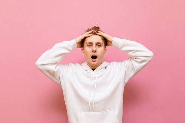 Şok olmuş, sıradan giyinmiş genç bir adam pembe bir arka planda duruyor, kafasını tutuyor, şaşırmış bir yüzle kameraya bakıyor. Yakışıklı adam şok oldu. Boşluğu kopyala