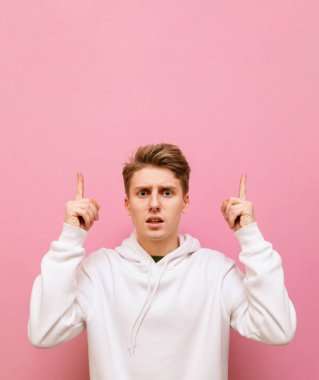 Beyaz kapüşonlu şaşkın adam pembe arka planda izole edilmiş, kameraya bakıyor ve fotokopi çekiliyor. Şok olmuş genç bir adamın boş bir yeri işaret eden dikey portresi