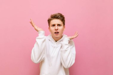 Beyaz kapüşonlu kafası karışmış genç adam pembe arka planda duruyor ve elleri havada şüpheli bir şekilde kameraya bakıyor. Pembe arka planda izole edilmiş kafası karışmış bir adamın portresi. Boşluğu kopyala