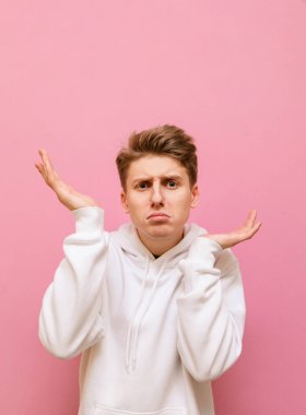 Beyaz kazaklı, şaşkın genç bir adam pembe arka planda duruyor, kollarını yana doğru açıyor, kafasını karıştırarak kameraya bakıyor. Pembe arka planda izole edilmiş kafası karışık bir adam. Dikey
