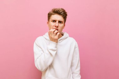 Pembe arka planda izole edilmiş beyaz kapüşonlu gergin genç adam endişeli bir bakışla kameraya bakıyor ve tırnaklarını yiyor. Pembe arka planda endişeli bir adamın portresi. Boşluğu kopyala