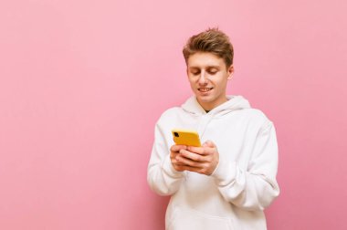 Pembe arka planda duran, elinde akıllı bir telefonla ekrana bakıp gülümseyen, günlük giysiler giyen genç bir adam. İzole edilmiş. Boşluğu kopyala