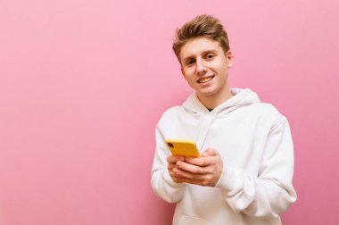 Beyaz kapüşonlu mutlu genç adam pembe arka planda, ellerinde Şirin 'le yalnız ve pozitif yüzlü kameraya bakarak gülümsüyor. Genç, pembe arka planda akıllı telefon kullanıyor. Boşluğu kopyala