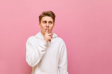 Açık renk giysili sevimli sarışın adam parmağıyla bir sessizlik hareketi gösteriyor ve yüzünde bir gülümsemeyle fotokopi odasına bakıyor. Neşeli genç adam pembe arka planda shsh jest yapıyor..