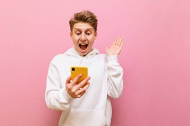 Akıllı telefon ekranına bakan ve elleri ve ağzı açık bir şekilde pembe arka planda izole edilmiş beyaz günlük kıyafetler giyerek sevinen genç adam şok oldu. Boşluğu kopyala