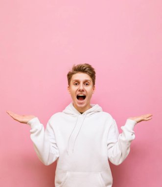 Pembe arka planda beyaz kapüşonlu, şaşırmış genç bir adamın portresi. Şok olmuş yüzüyle kameraya bakıyor ve ellerini yana açıyor. İzole edilmiş. Dikey.