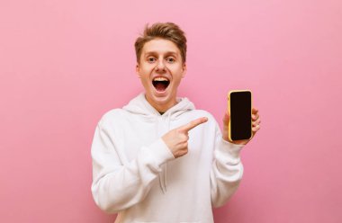 Pembe arka planda duran mutlu genç adam pembe arka planda elinde telefonuyla kameraya bakıyor ve siyah ekranda parmağını gösteriyor. Boşluğu kopyala