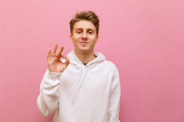 Beyaz gündelik giysiler içindeki yakışıklı genç adam yüzünde bir gülümsemeyle kameraya bakar ve el hareketi yapar. Güzel bir jesti olan yakışıklı sarışın adam pembe arka planda izole edilmiş. Boşluğu kopyala