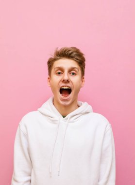 Pembe arka planda beyaz kapüşonlu mutlu adamın dikey portresi neşeli yüzüyle kameraya bakıyor ve ağzını açıyor. Pembe üzerine izole edilmiş neşeli genç bir adam. Boşluğu kopyala