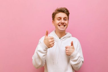 Neşeli genç sarışın adam kameraya bakıyor ve pembe arka planda izole edilmiş yüzünde bir gülümsemeyle baş parmaklarını gösteriyor. Pozitif adam jest yapar ve sevinir. Boşluğu kopyala