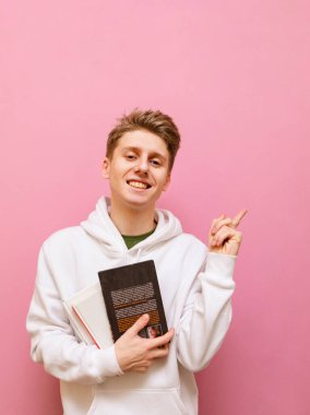 Pembe arka planda izole edilmiş, elinde kitaplarla mutlu bir genç portresi. Yüzünde gülümseme ve ellerinde kitaplarla işaret parmağını gösteren bir öğrenci.