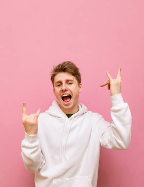 Beyaz giysili, pozitif genç bir adam kaya jesti sergiliyor ve mutlu bir yüzle kameraya bakıyor. Sarışın adam pembe arka planda ağır metal hareketi gösteriyor. Dikey.