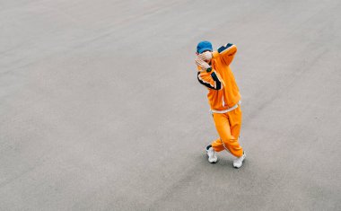 Asfalt bir meydanda hip hop dansı yapan şık genç adam, turuncu kıyafetler giyiyor. Modaya uygun sokak dansçısı gri asfalt arka planda hip hop performansı gösteriyor. Uzayı kopyala Arkaplan.