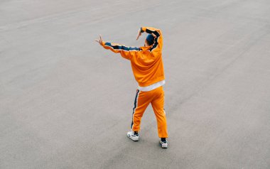 Modaya uygun bir sokak dansçısı, arka planda gri asfalt üzerinde hip hop performansı sergiliyor, ev hareketleri yapıyor. Turuncu eşofmanlı adam asfalt kaldırımda hip hop dansı yapıyor. Arkaplan