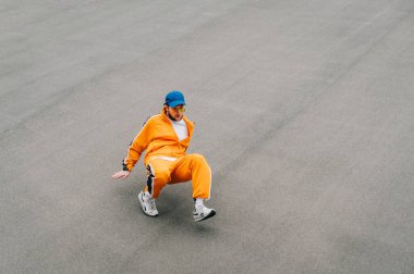 Parlak elbiseli bir adam asfaltta dans ediyor. Profesyonel hip hop dansçısı, turuncu giysiler içinde asfalt parkın arka planında bir performans sergiliyor. İzole edilmiş. Boşluğu kopyala
