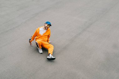 Mutlu hip hop dansçısının şık, parlak sokak kıyafetleriyle asfaltlanmış çocuk parkında oturduğu fotoğraf. Gri asfalt arka planda izole bir şekilde dans eden neşeli genç adam. Geçmişi. Boşluğu kopyala