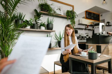 Resmi giysiler içinde genç bir iş kızı portresi. Elinde bir fincan kahve ve bir laptop ile rahat bir kafede oturuyor. Elinde iş evrakları var.