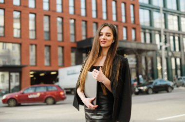 Karanlık giyinmiş genç bir iş kadını elinde dizüstü bilgisayarla sokaklarda poz veriyor, yüzünde bir tebessümle kameraya bakıyor, kablosuz kulaklık takıyor. İş adamları.