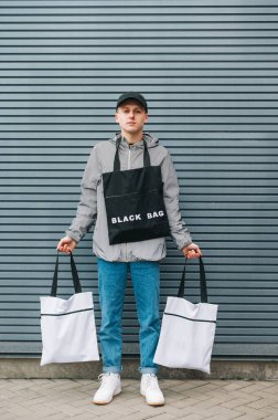 Şık giyinmiş yakışıklı bir gencin portresi gri bir duvarın arka planında elinde ekolojik poşetlerle dikey boyda bir portre ile kameraya poz veriyor. Çevre dostu adam.