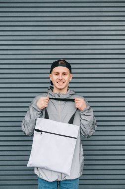 Gri bir arka planda elinde beyaz bir alışveriş çantasıyla şık giyinmiş gülümseyen genç adam yüzünde bir gülümsemeyle kameraya bakıyor. Çevre dostu adam çevre çantasıyla poz veriyor..