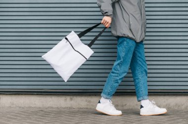 Gri bir duvarın arka planında elinde beyaz bir çevre çantasıyla gezen şık elbiseli bir gencin kesilmiş fotoğrafı. Çevre dostu. Delikanlının elinde yeniden kullanılabilir çanta. Boşluğu kopyala