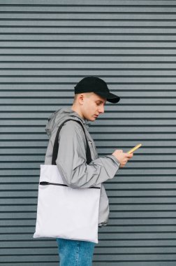 Şık sokak kıyafetleri içindeki genç adam omuzunda alışveriş çantasıyla duruyor ve ciddi görünümlü bir akıllı telefon kullanıyor. Çevre dostu genç adam interneti akıllı telefonda kullanıyor. Dikey fotoğraf.