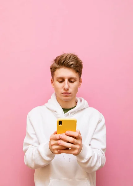 Portrait Concentrated Young Man White Hoodie Stands Pink Background Smartphone — Stock Photo, Image