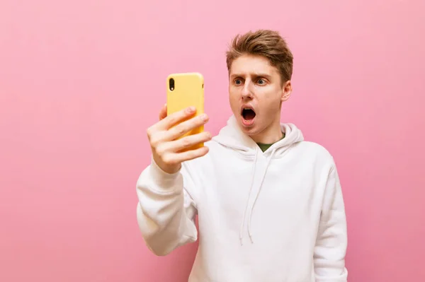 Shocked Young Man White Sweatshirt Stands Smartphone Hand Pink Background — Stock Photo, Image