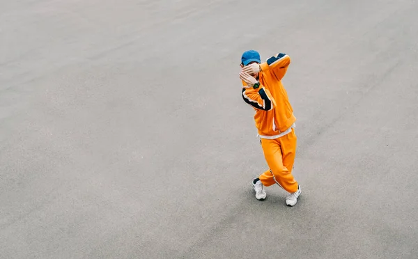 Stylish young man dancing hip hop on an asphalt square, wearing orange clothes. Fashionable street dancer shows hip hop performance on gray asphalt background. Copy space. Background.