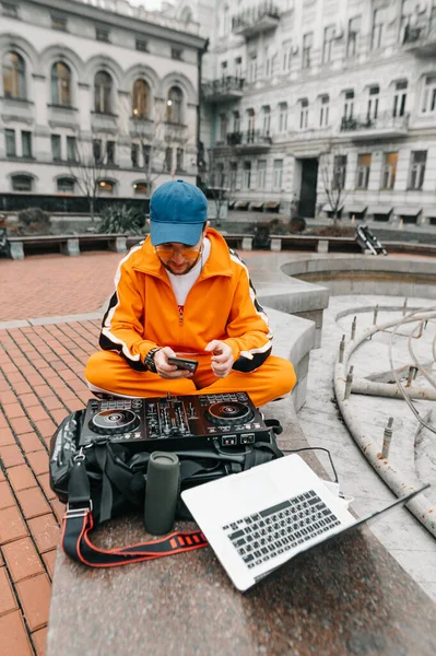 一个穿着明亮休闲装的Dj坐在外面 带着笔记本电脑和控制器 播放音乐 并使用智能手机 一个街头Dj和一个在街上放音乐的混音器的照片 — 图库照片