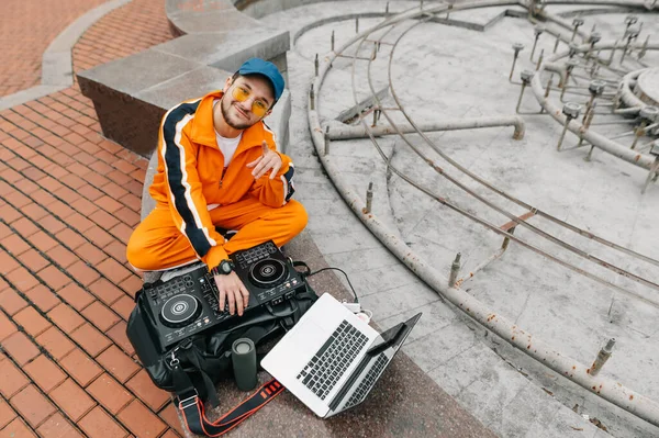 Wesoły Człowiek Pomarańczowych Ubraniach Okularach Siedzi Kontrolerem Muzyki Laptopem Ulicy — Zdjęcie stockowe